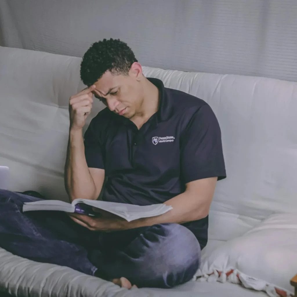 A man sits on a couch with a book open on his lap, looking thoughtful—perhaps pondering, "Is a degree needed to become a game developer?" A laptop is beside him, and he is wearing a dark polo shirt and jeans.