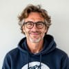 Middle-aged man with wavy hair, glasses, and a beard, smiling while wearing a navy blue hoodie—he stands against a plain light background, embodying the approachable spirit often seen in About Game Developers profiles.